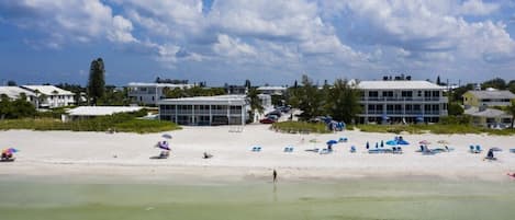 A beachfront hotel with multiple buildings, a sandy beach with several umbrellas and lounge chairs, and a clear sky.