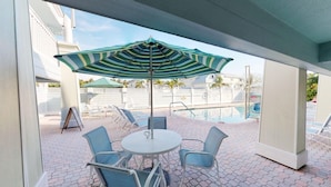 A covered outdoor seating area with a table and chairs, a striped umbrella, and a pool in the background. - Holmes Beach | Couples Gulf Coast Retreat | Pool + Beach (Holmes Beach)