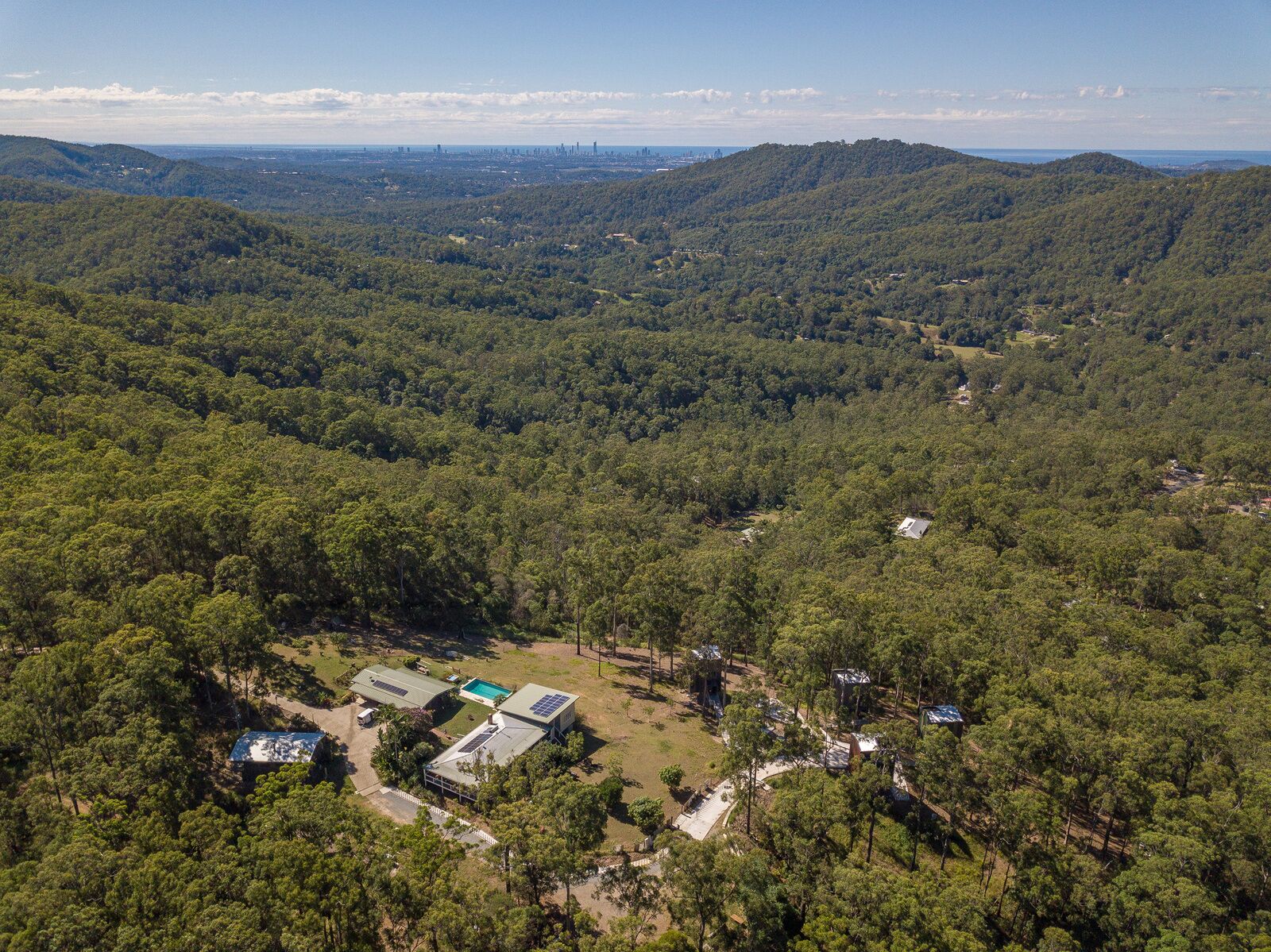 An aerial view of a forested area with a small cluster of buildings, including a house with solar panels and a swimming pool.