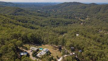 An aerial view of a forested area with a small cluster of buildings, including a house with solar panels and a swimming pool.