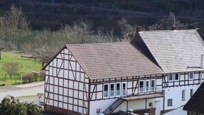 A traditional half-timbered house with a steeply pitched roof, a visible staircase, and a view of a green landscape in the background.