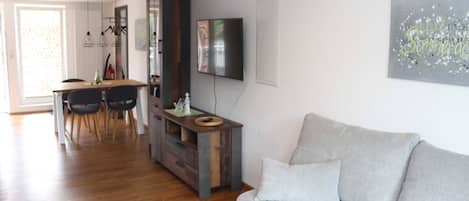 Una sala de estar con sofá gris, piso de madera, zona de comedor con mesa y sillas, y un televisor montado en la pared.
