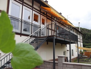 Un edificio de dos pisos con balcón, escaleras y un patio con mobiliario exterior.