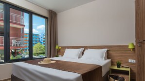 A hotel room with a large bed, a small bedside table with a lamp, a window with a view of buildings, and a wooden headboard.