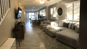 A modern living room with a sectional sofa, a flat-screen TV mounted on the wall, a dining area with a bar, and a kitchen in the background. - Modern Remodel + Amenities Galore! (Gilbert)