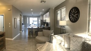 A modern living room with a sofa, a dining area with chairs, a kitchen in the background, and a large wall clock. - Modern Remodel + Amenities Galore! (Gilbert)