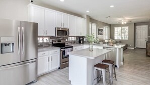 A modern kitchen with white cabinets, stainless steel appliances, and a central island with stools. - Modern Remodel + Amenities Galore! (Gilbert)