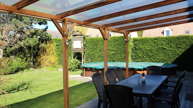 A covered patio with a wooden deck, outdoor furniture, and a view of a well-maintained garden and neighboring buildings. - Maison du Lavoir Disneyland (Bailly-Romainvilliers)