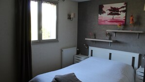 A bedroom with a bed, a window with curtains, a radiator, a nightstand, a wall-mounted shelf with decorative items, and a painting on the wall. - Maison du Lavoir Disneyland (Bailly-Romainvilliers)