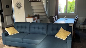 A modern living room with a blue sectional sofa, a wooden coffee table, a dining table with chairs, a staircase, and a large window with a view of greenery. - Maison du Lavoir Disneyland (Bailly-Romainvilliers)