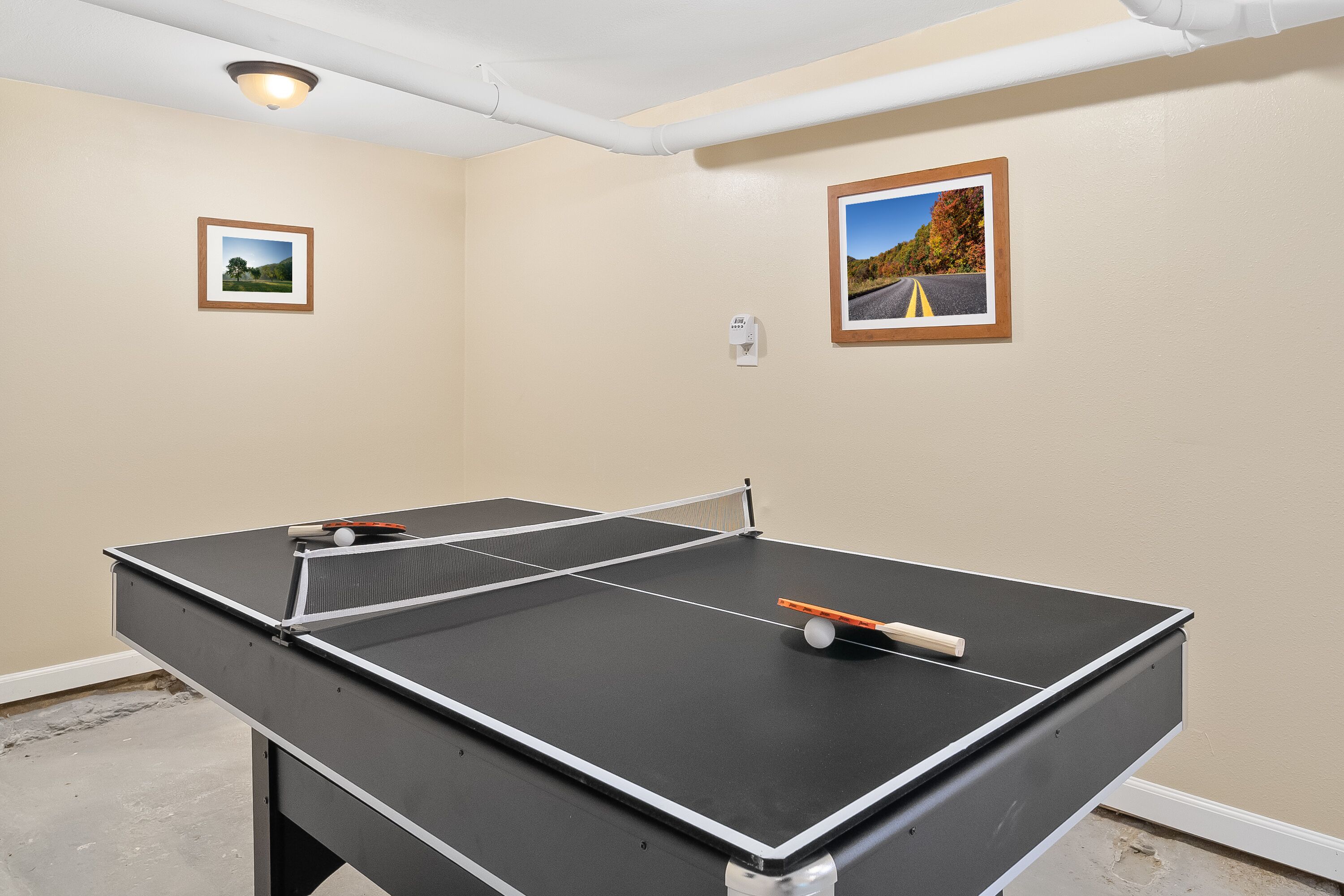 A ping pong table in a room with beige walls, two framed pictures, and a ceiling light fixture.