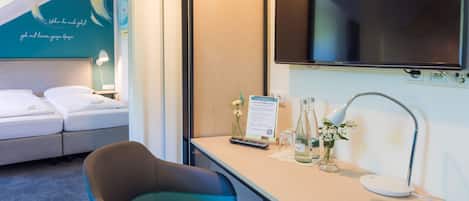 A hotel room with a bed, a desk with a lamp, a TV, and a sign welcoming guests.