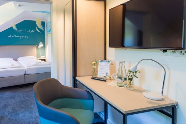 A hotel room with a bed, a desk with a lamp, a TV, and a sign welcoming guests. - Hotel & Café Mühlenglück (Oppenau)