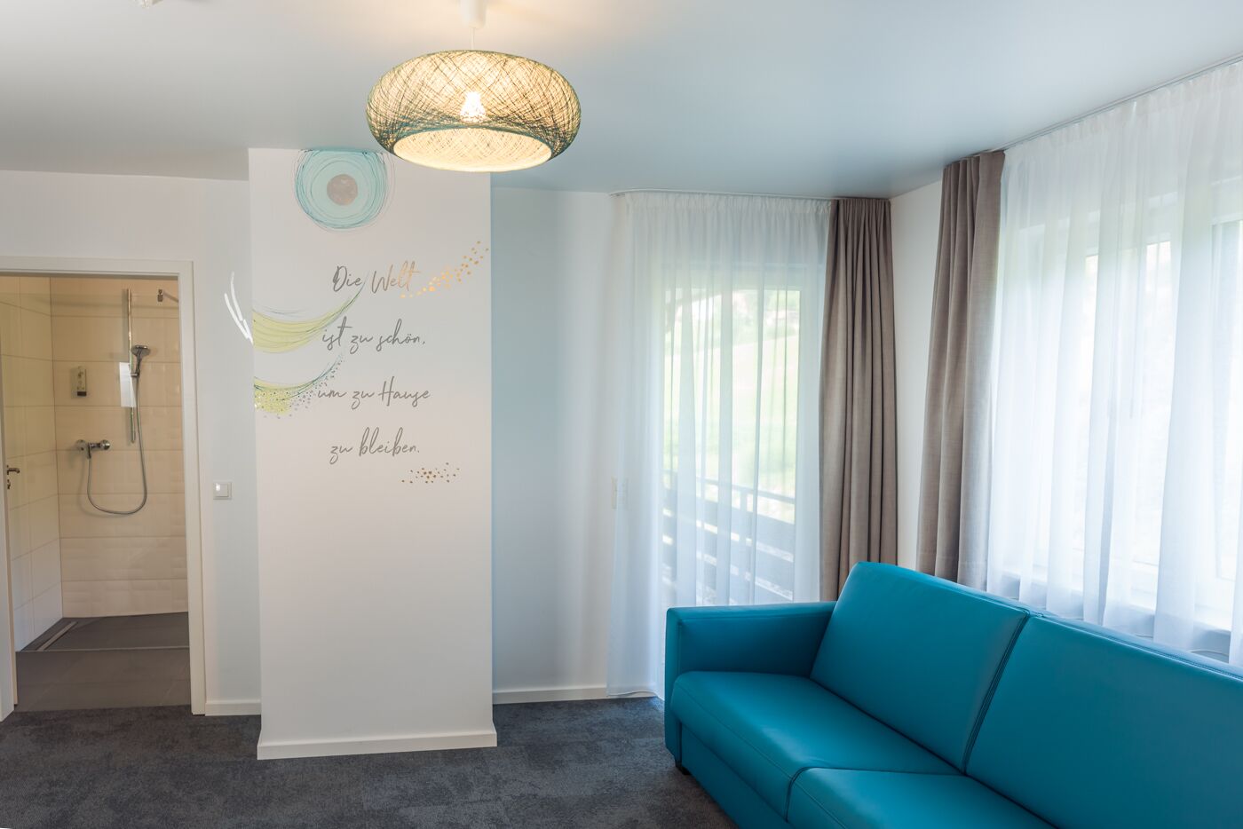 A modern living room with a teal sofa, a decorative wall with text, a shower area, and sheer curtains.