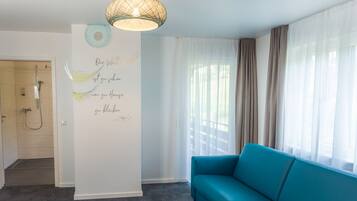 A modern living room with a teal sofa, a decorative wall with text, a shower area, and sheer curtains.