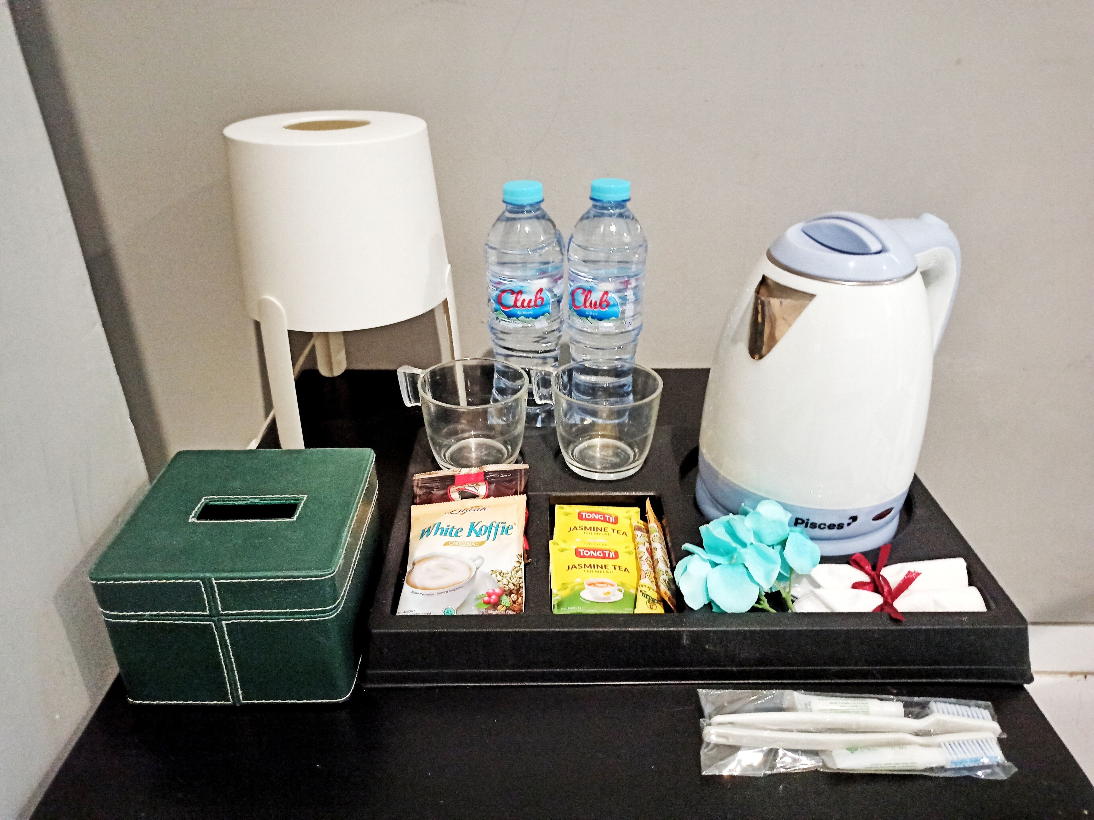 Un mini-bar d’hôtel comprenant une boîte verte, deux bouteilles d’eau de la marque Club, une bouilloire, une machine à café, une boîte de sachets de thé, une brosse à dents et un rouleau de mouchoirs.