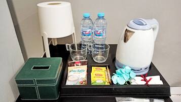 Un mini-bar d’hôtel comprenant une boîte verte, deux bouteilles d’eau de la marque Club, une bouilloire, une machine à café, une boîte de sachets de thé, une brosse à dents et un rouleau de mouchoirs.