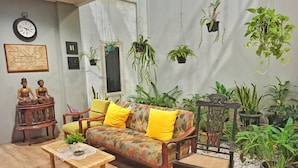 A living room with a floral sofa, yellow cushions, a wooden coffee table, and various hanging plants. - Manggolo Homestay Syariah (Yogyakarta)