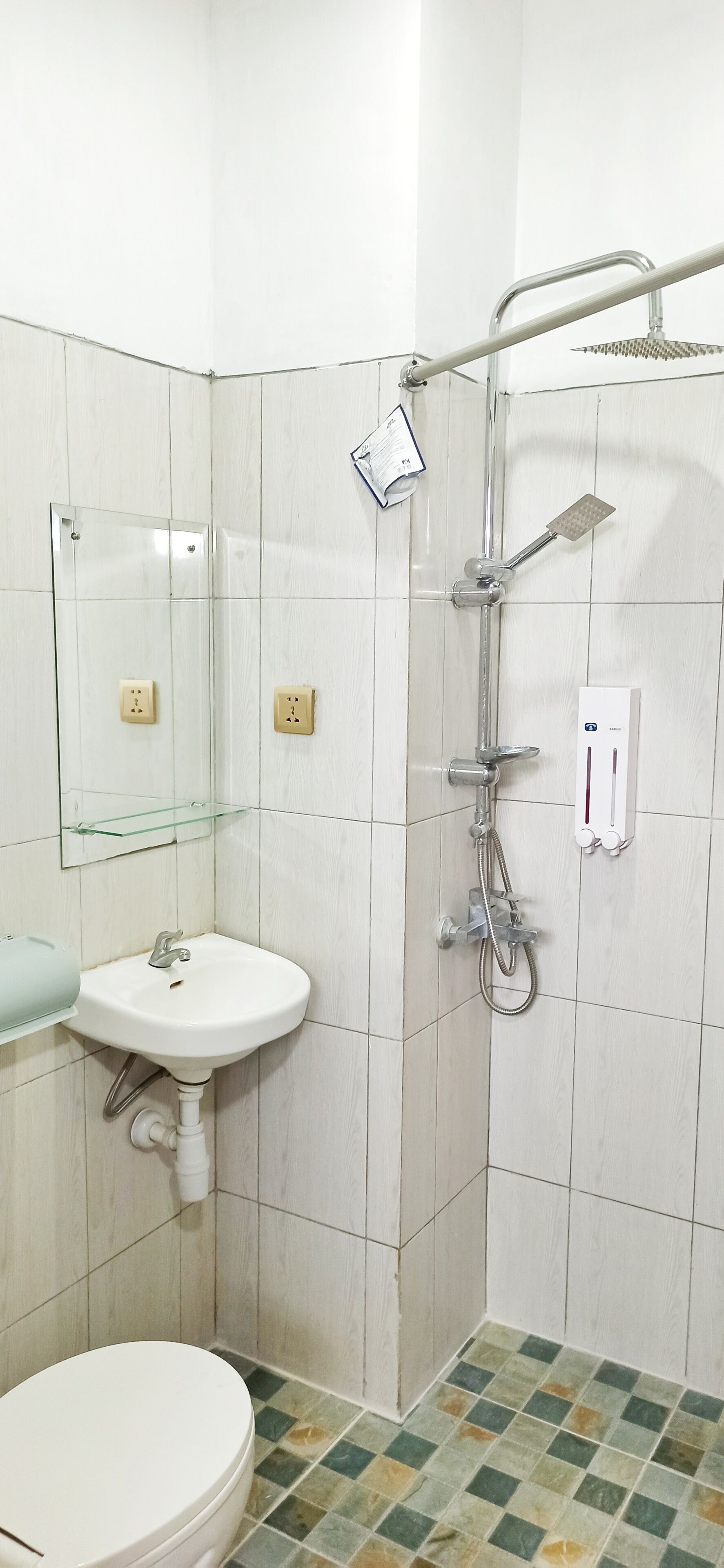 Une salle de bain avec un lavabo blanc, une étagère en verre, des toilettes et une douche équipée d’un pommeau de douche à main.