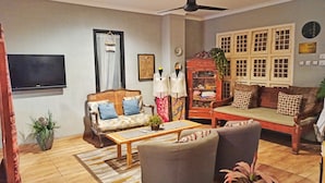 A living room with a sofa, armchairs, a coffee table, a television, a ceiling fan, and a cabinet with glass doors. - Manggolo Homestay Syariah (Yogyakarta)