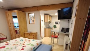 Een compacte slaapkamer met een bed, een klein bureau, een televisie en een kitchenette op de achtergrond.
