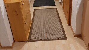 Een smalle gang met houten kasten, een zwarte mat bij de ingang, en een deur met een ruit.