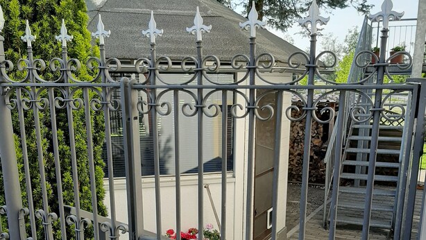Een omheind gebied met een klein gebouwtje, mogelijk een schuurtje of opslagruimte, omringd door een metalen hekwerk met decoratieve details.