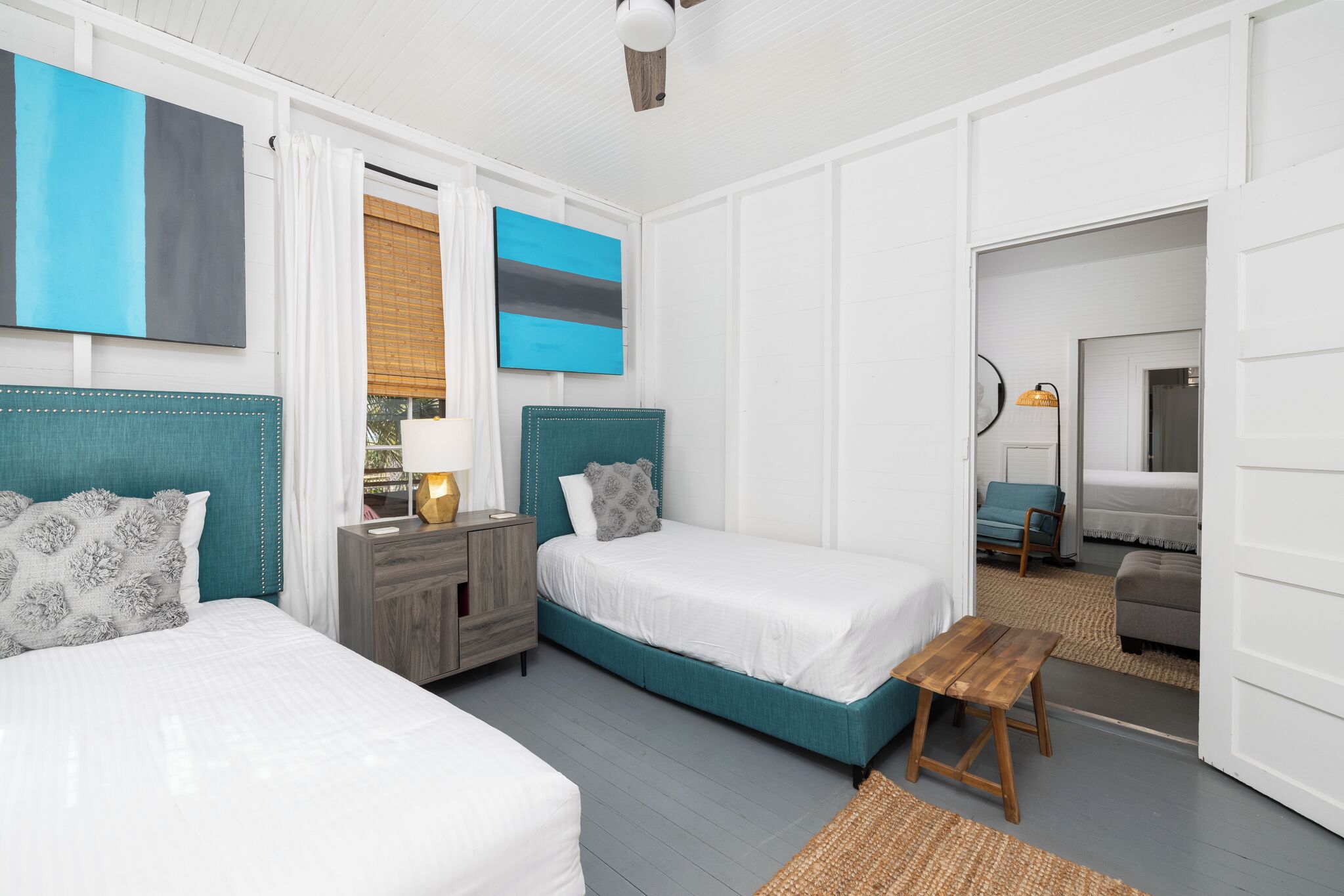 A bedroom with two beds, a nightstand, a wooden bench, and a view of another room.