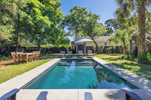 A rectangular swimming pool with a clear blue water surface, surrounded by a concrete deck. There are four lounge chairs aligned along the pool's edge, and a wooden table with chairs is positioned to the left.