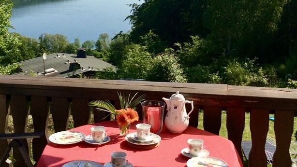 A table set with tea cups, plates, and a teapot on a red tablecloth, overlooking a lake with a view of a town and hills in the distance. - House with lake view and terraces - spacious (Edertal - Bringhausen)