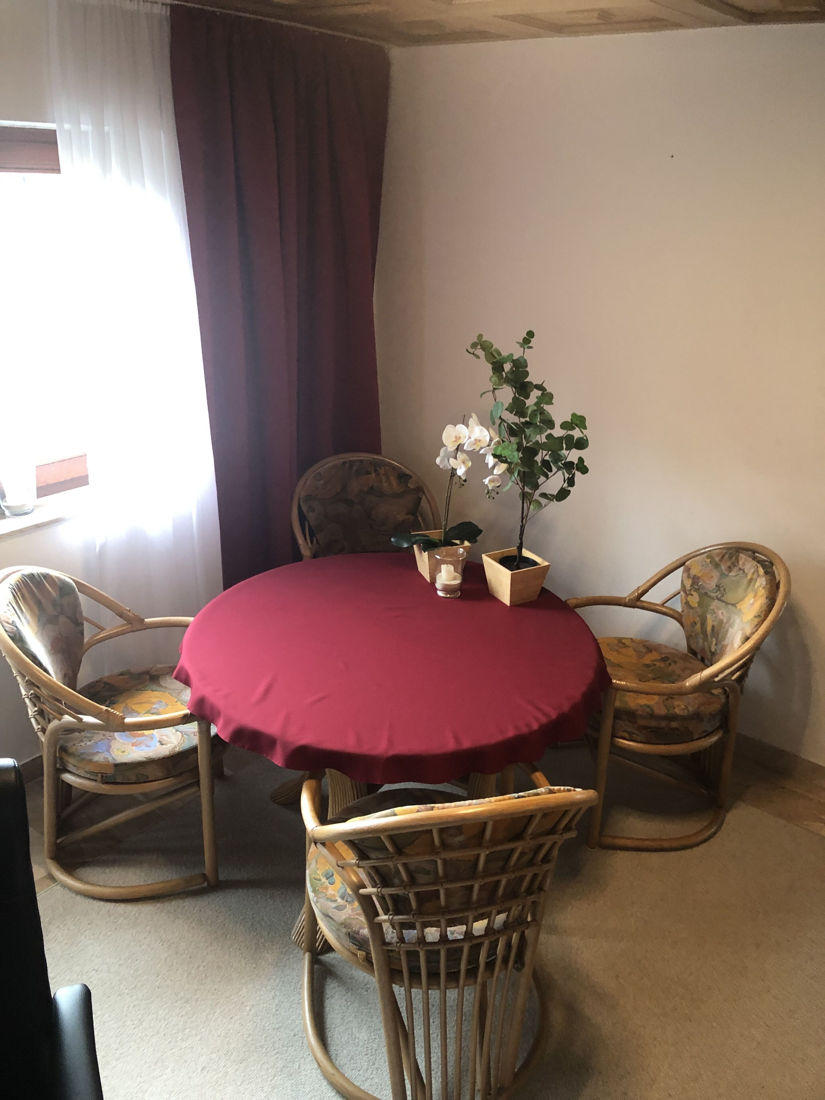 Una mesa redonda con mantel rojo rodeada de cuatro sillas de mimbre. Hay una orquídea en maceta sobre la mesa.