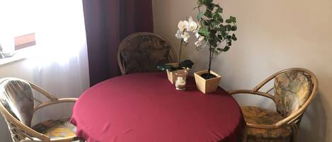 Una mesa redonda con mantel rojo rodeada de cuatro sillas de mimbre. Hay una orquídea en maceta sobre la mesa.