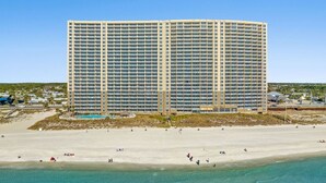 Ein großes, mehrstöckiges Hotel mit flachem Dach und zahlreichen Balkonen, direkt an einem Strand mit klarem, blauem Wasser gelegen.