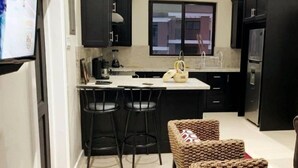 Una cocina moderna con gabinetes negros, una encimera blanca y un ventanal. Hay dos taburetes negros y una silla de mimbre con un cojín rojo.
