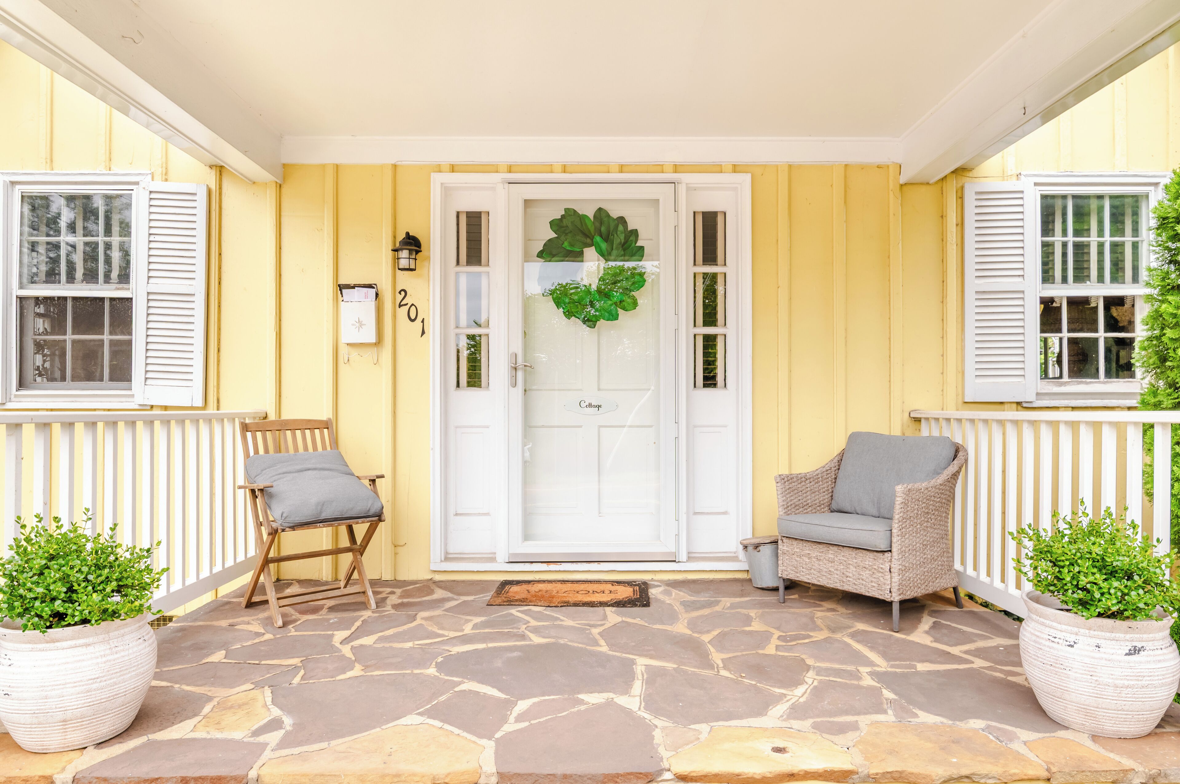 Un bâtiment jaune avec une porte blanche, deux chaises en osier et des plantes en pot.