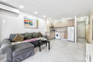 A modern apartment with a grey sofa, a coffee table, a washing machine, a microwave, and a refrigerator in the kitchen area.
