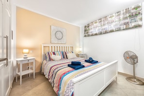 Une chambre à coucher comprenant un lit, des tables de chevet, une œuvre d’art accrochée au mur et un ventilateur sur pied.