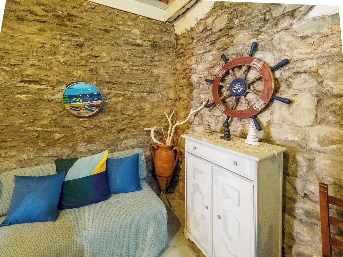 A room with a stone wall, a bed with blue and multicolored pillows, a white cabinet, and a ship's wheel decoration.