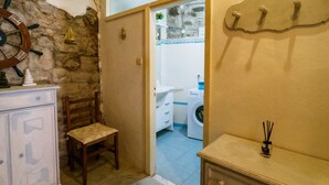 A room with a stone wall, a wooden chair, a white cabinet, and a view into a bathroom with a washing machine.