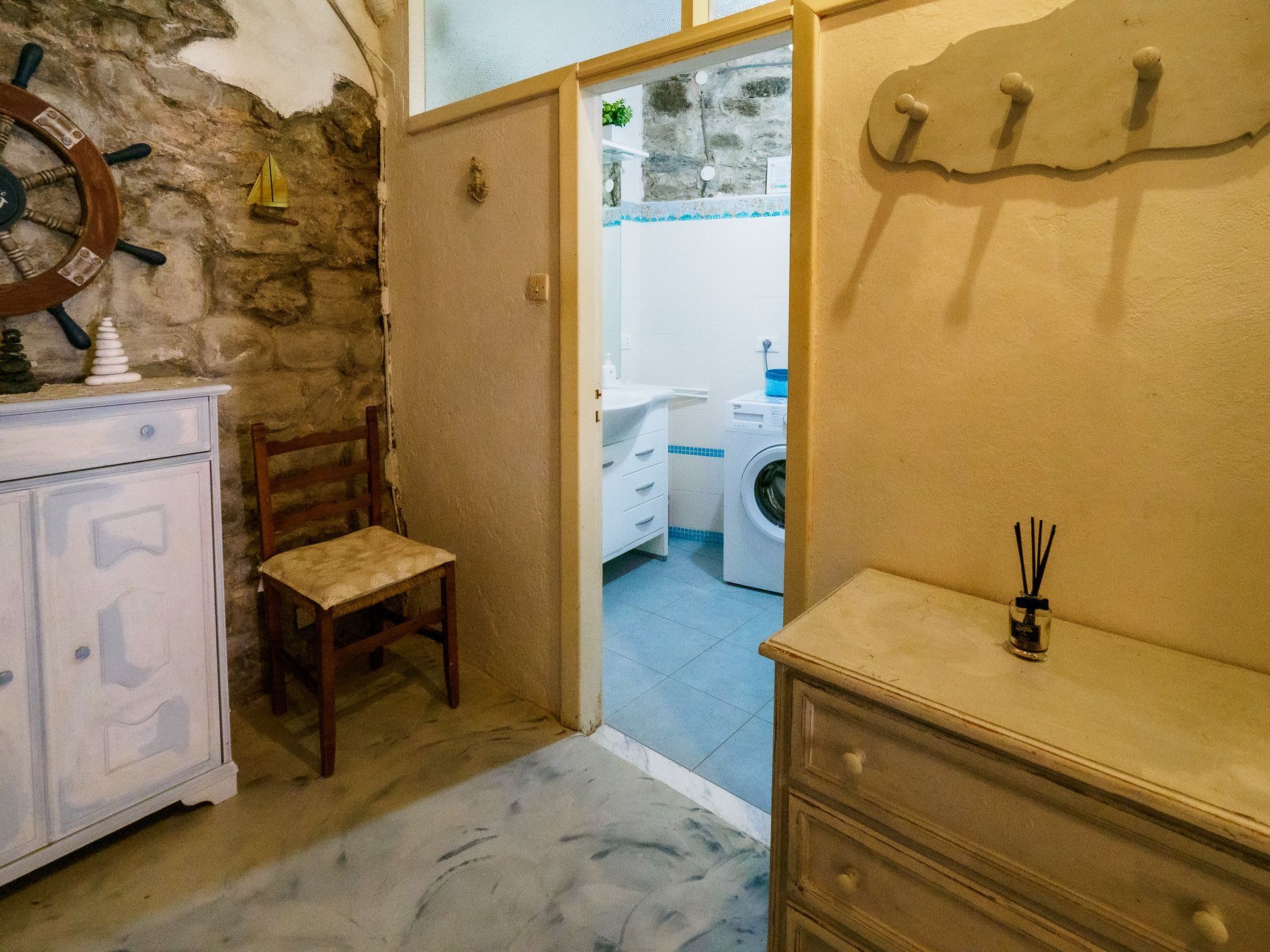 Quarto com parede de pedra, uma cadeira de madeira, um armário branco e uma porta de entrada para um banheiro com máquina de lavar.