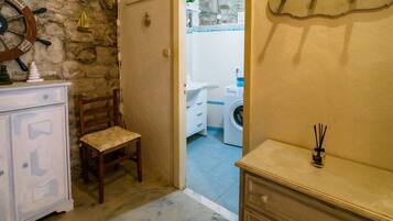 Quarto com parede de pedra, uma cadeira de madeira, um armário branco e uma porta de entrada para um banheiro com máquina de lavar.