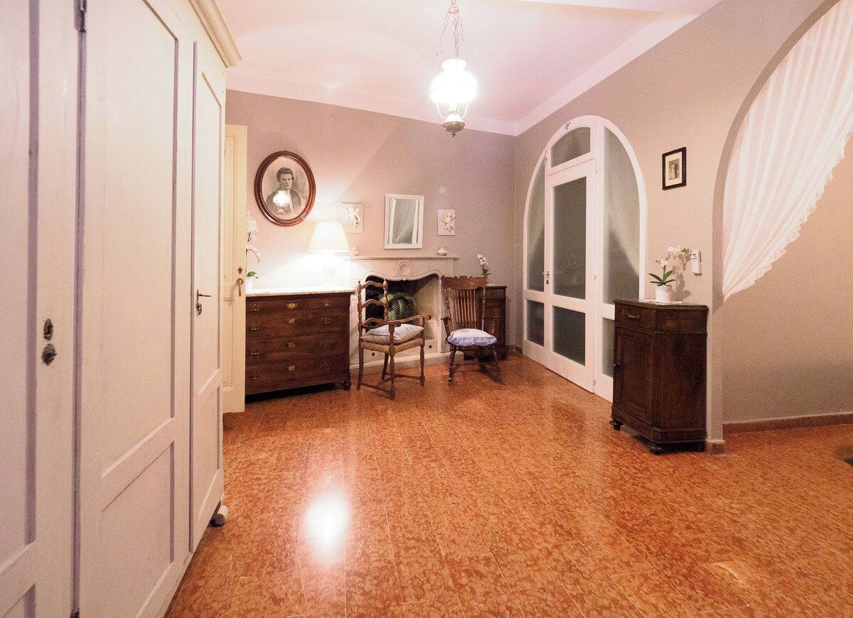 A room with a wooden floor, a desk with a lamp, a chair, a mirror, and a large arched window.