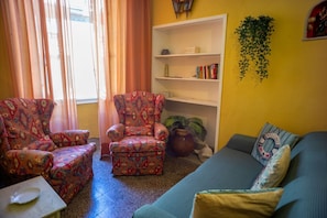 Un salon avec des fauteuils à motifs, une bibliothèque, un canapé et une fenêtre avec des rideaux.