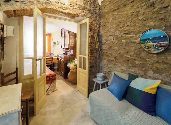 A rustic room with a stone wall, a sofa with cushions, a dining table, and a kitchen area visible through an open door.