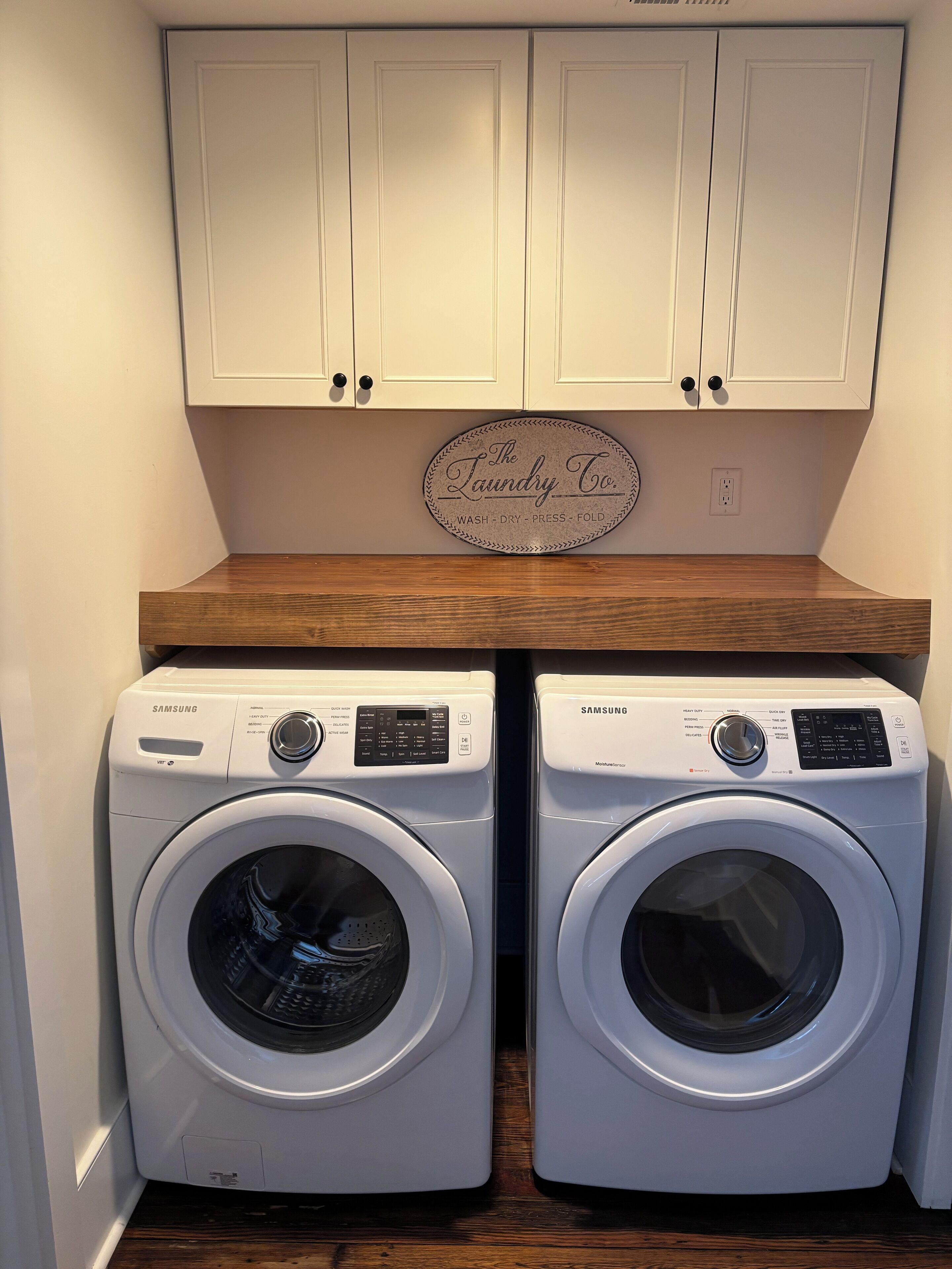 Two Samsung front-loading washing machines in a laundry room with white cabinets above and a wooden countertop below.