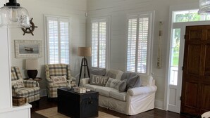 Un salon spacieux avec un canapé blanc, un fauteuil à motifs et une table basse. On y trouve un ventilateur de plafond, une lampe et une fenêtre avec des stores.