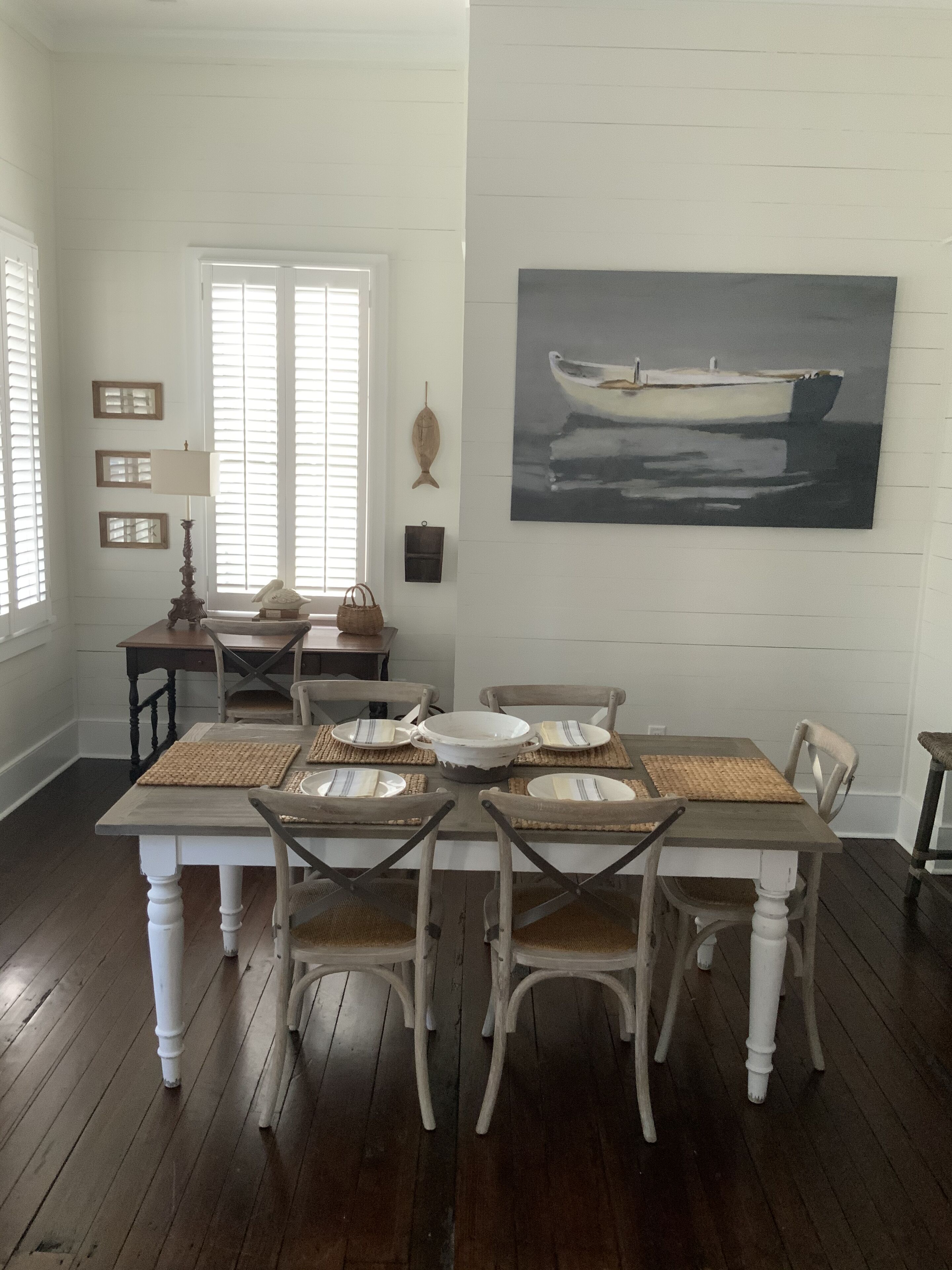 Uma sala de jantar com uma mesa e cadeiras de madeira, um quadro de um barco na parede e uma janela com persianas.