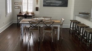 Un coin repas avec une table en bois, des chaises blanches et un tableau représentant un bateau accroché au mur.
