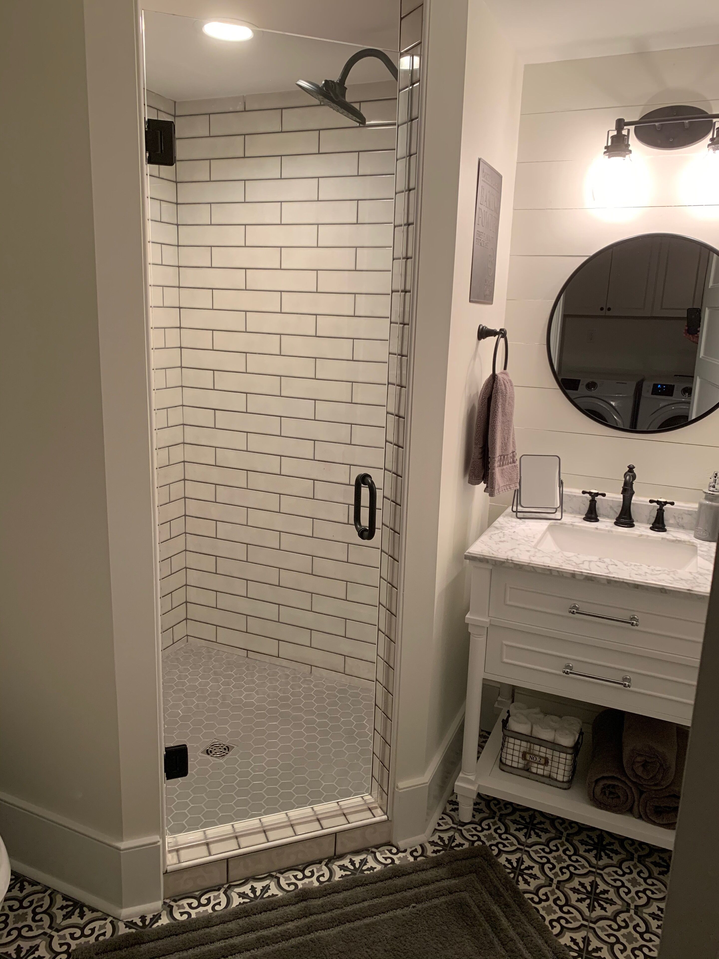Une salle de bain dotée d’une douche à parois de verre, d’un meuble-lavabo blanc avec un plan de travail en marbre et d’un miroir rond au-dessus du lavabo.