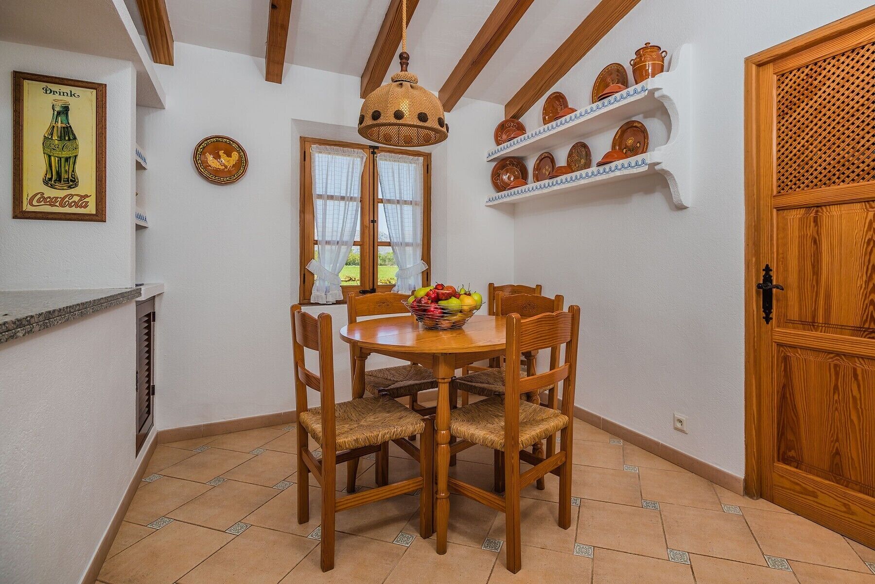 A dining area with a wooden table and chairs, a fruit bowl, a woven basket hanging from the ceiling, and shelves with decorative plates.